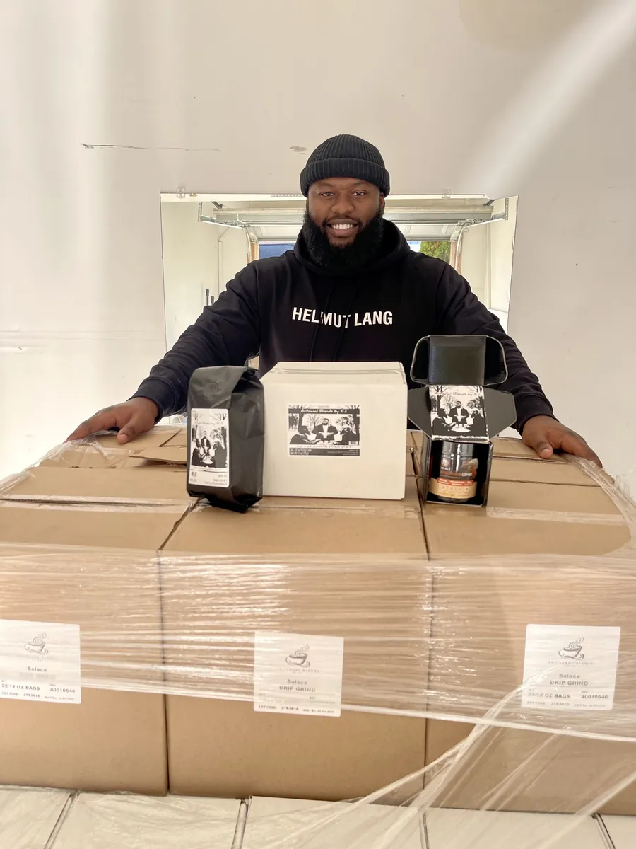 Emmanuel Etoo smiling behind a wrapped pallet of Solace coffee cases, with a bag, a box and packaging from the first shipment